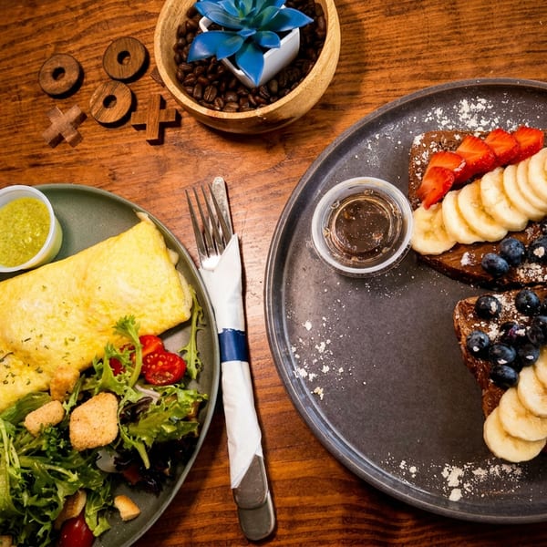 Overhead view of an omelet with salad, fruit-topped French toast, dipping sauces, and a succulent on a wooden restaurant table.