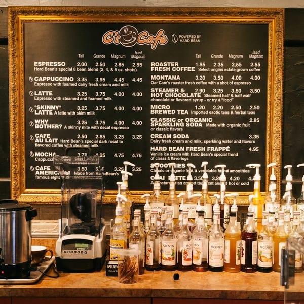 Coffee counter featuring a framed café menu, espresso equipment, blender, beverage dispensers, and numerous flavored syrup bottles.