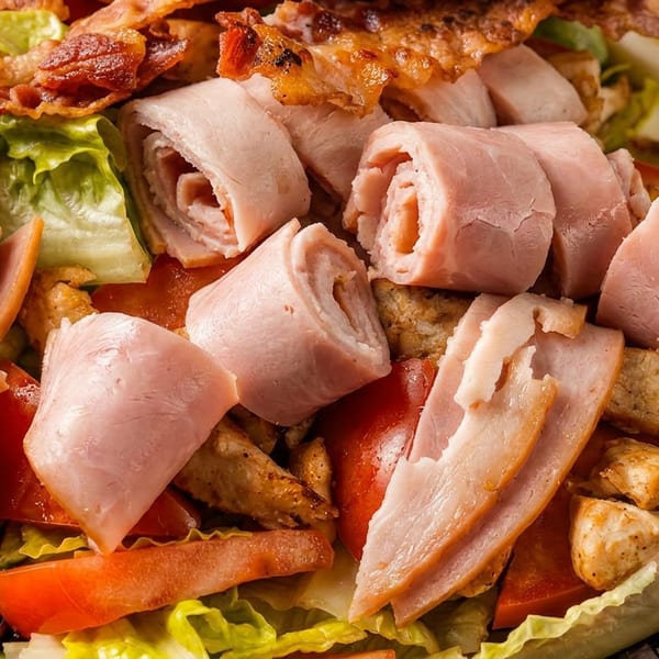 Close-up of a hearty salad topped with rolled ham, crispy bacon, grilled chicken, tomatoes, lettuce, and sliced hard-boiled eggs.