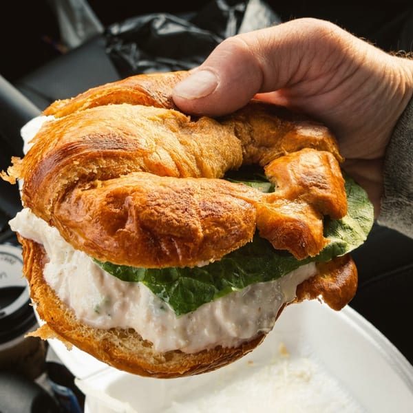 Hand holding a croissant sandwich filled with creamy salad and lettuce inside a car.