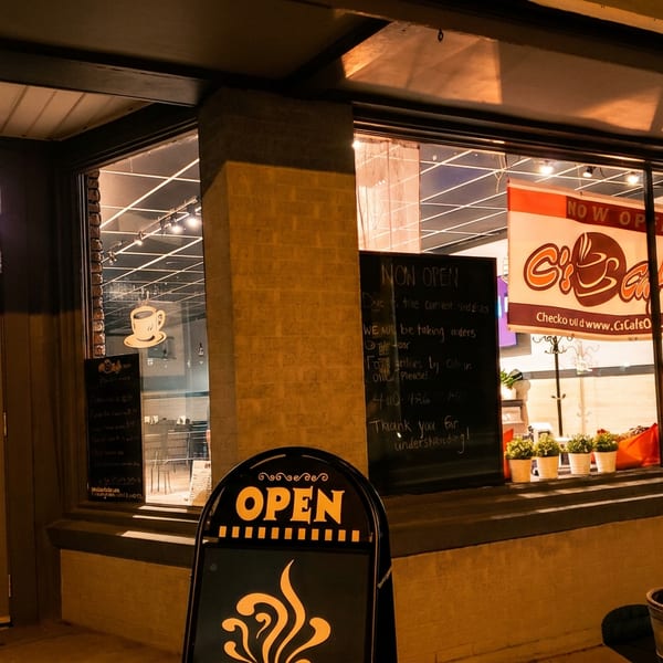 warmly lit café storefront at night displays open signs, a chalkboard message, potted plants, and a café banner.