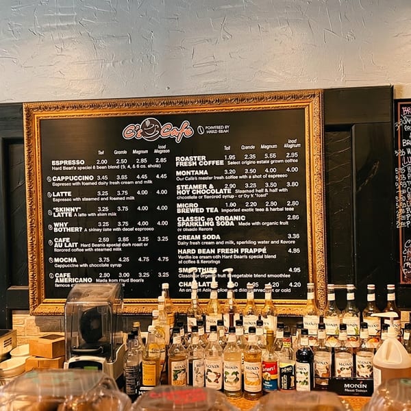 Cafe counter displaying coffee menu boards, syrup bottles, coffee equipment, and pastry containers.
