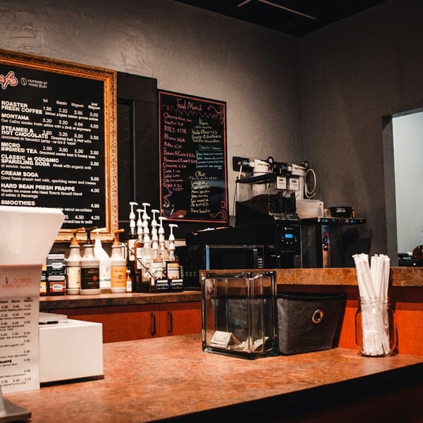 Cozy café counter with coffee menu, espresso machine, stacked cups, syrup bottles, straws, and register.